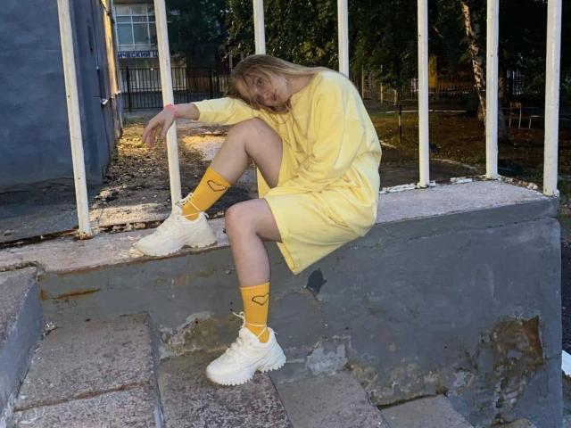 ShawtyMiaAndJack sits on concrete steps, wearing a yellow dress, yellow heart-patterned socks, and white sneakers, with a tired expression, arms resting on knees.