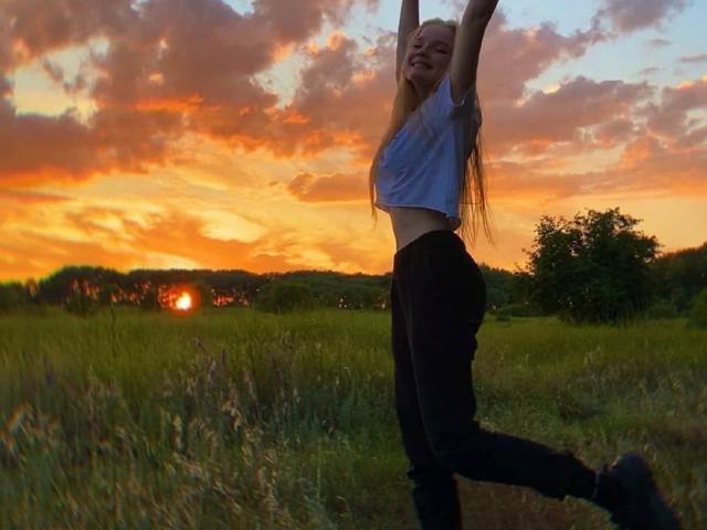 ShawtyMiaAndJack has long blonde hair, wearing a white crop top and black pants, stretching arms overhead in a grassy field at sunset, with a happy expression.
