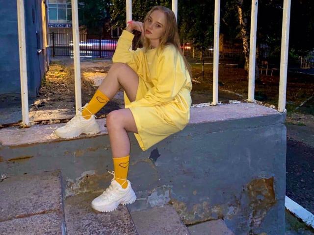 ShawtyMiaAndJack with long blonde hair, yellow oversized dress, yellow socks with heart, and white sneakers, sitting on concrete steps, right knee up, left hand near face.