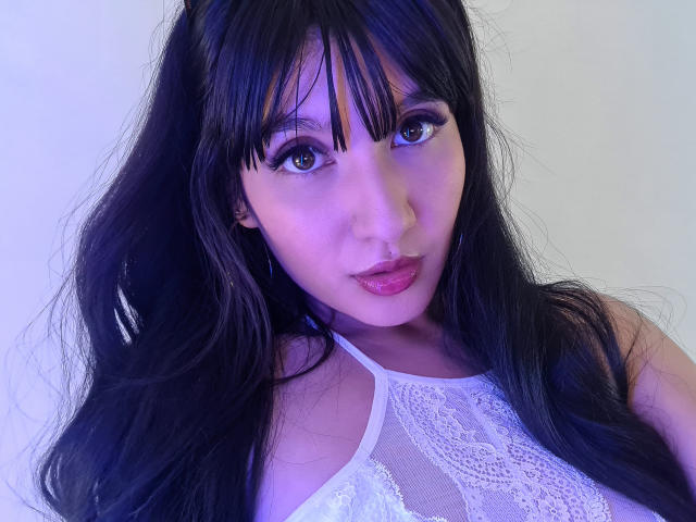 NaylaGrey, con cabello negro largo, flequillo y un tono púrpura-azul, vestida con una blusa blanca de encaje, mostrando sus labios rosados y sus ojos marrones, mirando directamente a la cámara..