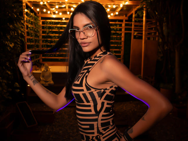 VicktoriaSunshine with long black hair, glasses, and a tan and black geometric dress, holding her hair, showing tattoos, and a confident smile, in a dimly lit, warm setting.