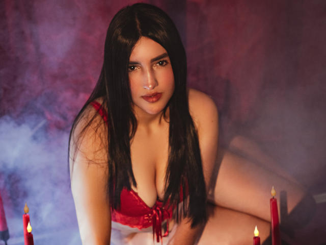 YviTaylor with long black hair, red lace bra, and red candles in the foreground, kneeling, looking at the camera, her tits slightly visible, with a smoky purple background.