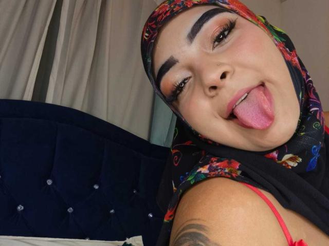 Close-up of InayaKhalifa. She has a light brown complexion, thick black eyebrows, and wears a colorful floral hijab. Her tongue is out, and she's looking at the camera with a playful expression. She has a small nose stud on her left nostril. Her red bra strap is visible. The background shows a dark blue, tufted headboard. Her tattoo is partially visible on her right upper arm. The image is well-lit, emphasizing her facial features and colorful hijab.