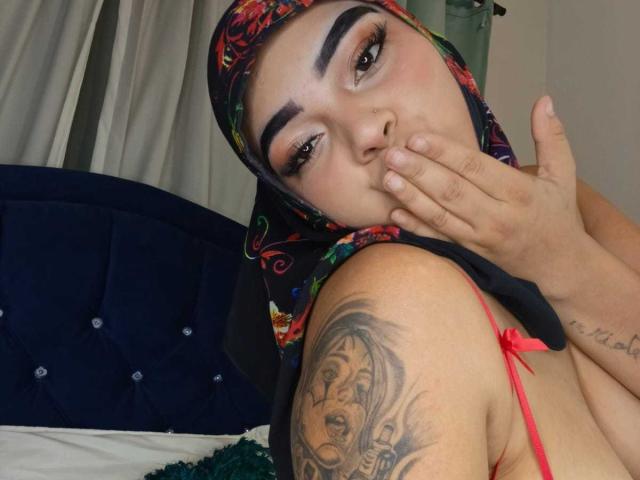 InayaKhalifa has a light brown skin tone and wears a colorful floral headscarf. She has thick, arched eyebrows, long eyelashes, and subtle pink blush. Her right hand covers her mouth. She has a tattoo of a woman's face on her right shoulder. She wears a red strap, revealing her bare shoulder. The blue velvet headboard is in the background. The image focuses on her facial expression and tattoo. The texture of her headscarf contrasts with her smooth skin.