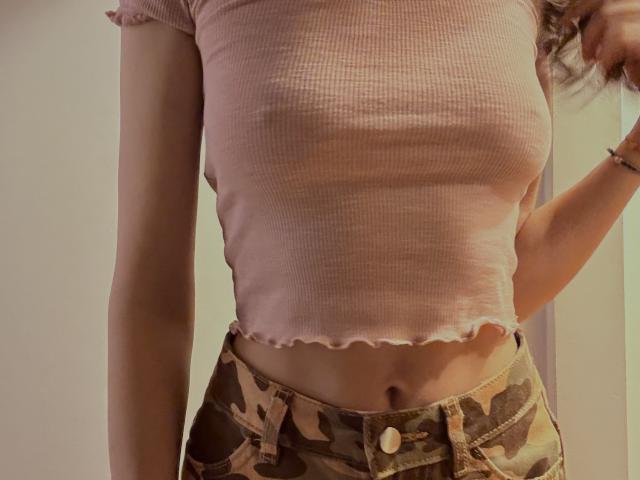 SweetClaraa porte un crop top rose texturé qui laisse légèrement entrevoir ses seins, et elle est vêtue d'un pantalon camouflage avec une boutonnière et un bracelet à son poignet droit..