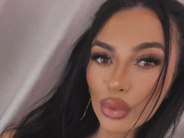 KinkyAmelie has dark hair, brown eyes, and full pink lips. She wears long gold earrings, and her makeup includes thick eyebrows and long lashes.