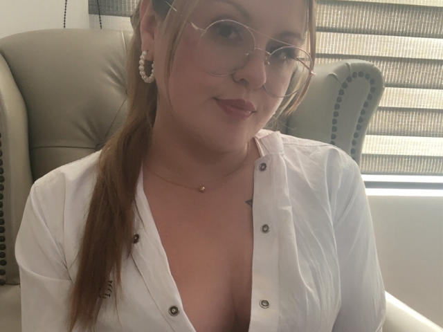 LaurenSpitz with glasses, white button-down shirt, visible cleavage, gold necklace, pearl earrings, brown hair in ponytail, sitting in office chair, smiling, light skin, subtle makeup.