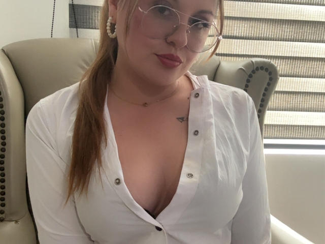 LaurenSpitz with glasses, white button-up shirt, deep cleavage, small tattoo on right chest, light skin, brown ponytail, subtle smile, sitting in beige office chair.