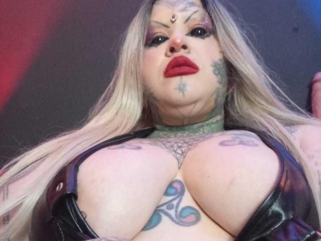 VenusHottt with long blonde hair, red lipstick, tattoos, large tits, green choker, black leather top, and a jewel on her forehead, looking down at the camera.