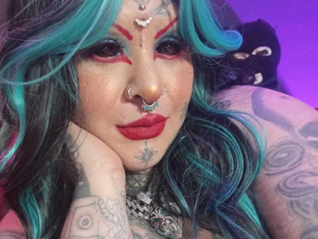 VenusHottt with teal and blue hair, nose piercings, red makeup, tattoos, red lipstick, and a necklace, leaning on her hand, showing her tattooed arm.
