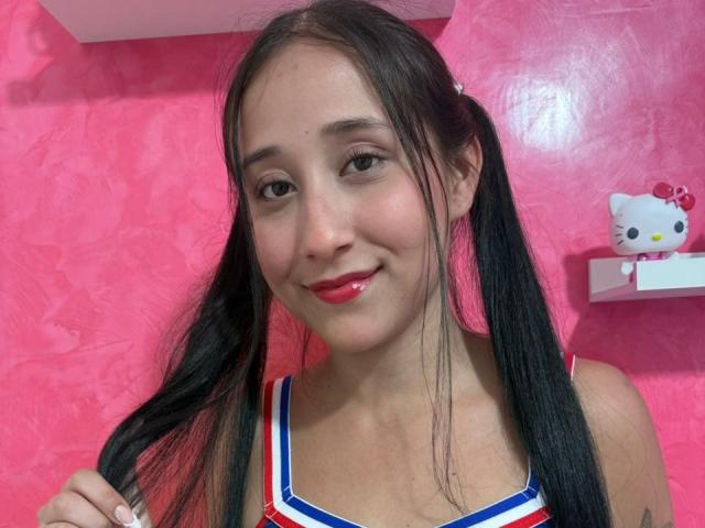 AnnieBlakke with long black pigtails, wearing a red and white cheerleader top, has red lipstick and is smiling gently, showing small tits against a pink wall.