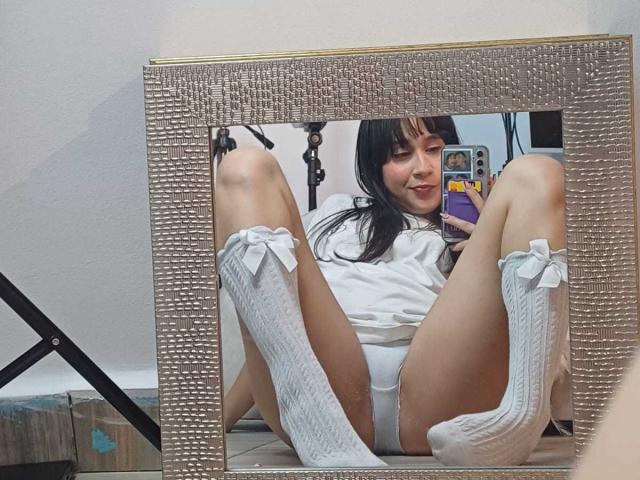 GlamorousLadies with black hair, white socks with bows, and white panties, sitting with legs spread, showing her pussy in a mirror. She's smiling, wearing a white shirt.