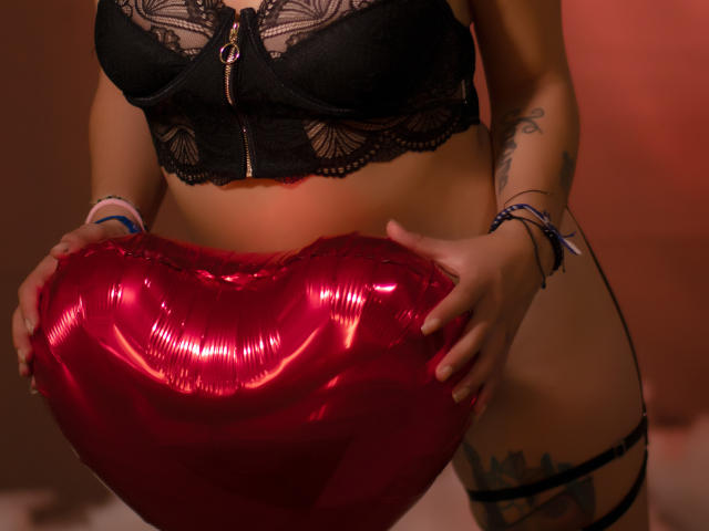 ValkiriaHolfs with big tits, wearing black lace bra, holding shiny red heart-shaped balloon, tattoos on right thigh, bracelets on wrists, soft red lighting.