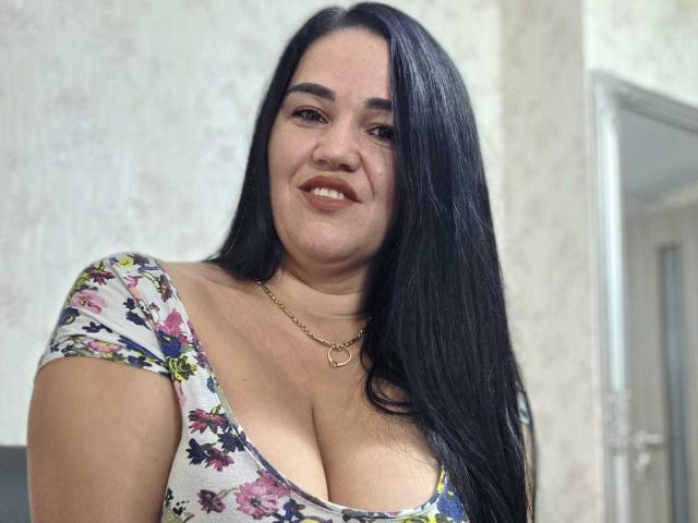 VickyDivinee has long black hair, tan skin, and a smile showing teeth. She's wearing a floral dress with a low neckline, showing her large tits and a gold necklace.