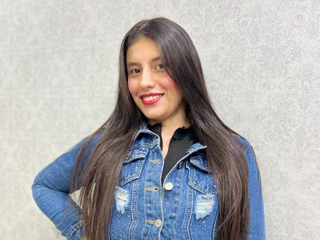 MadissonFontaine with long, straight black hair, blue denim jacket, black shirt, and red lipstick, smiling with hands on hips. Her tits are slightly visible through the jacket.
