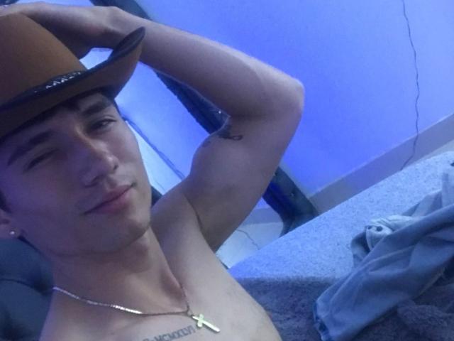 HombreMaloX, shirtless, wearing a brown hat, smiling, with a cross necklace, holding the hat, tattoo on right bicep, blue-lit room, grey blanket in background.