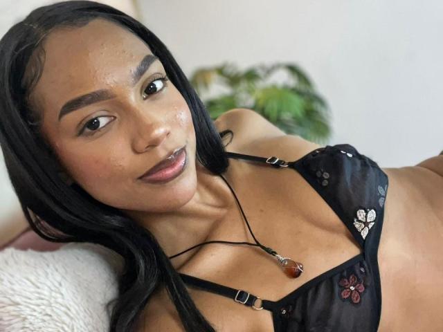 AylaCallowey with dark skin and black hair, wearing a black sheer bra with floral embroidery, showing her small tits and a red necklace, lying on a white pillow.