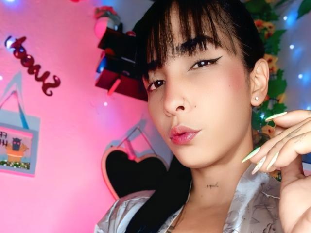 BrittanMelly with dark hair, bangs, and eyeliner, pouty lips, green nails, small tattoo on neck, wearing silky shirt, in pink and blue lit room.