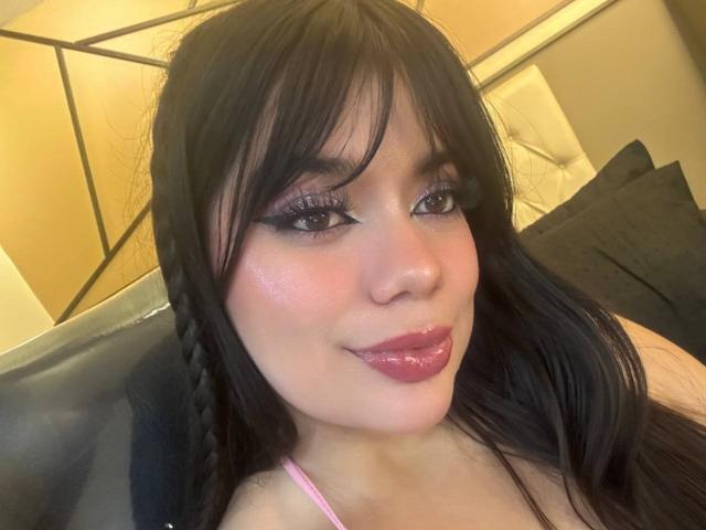 LeaMont with shiny pink lips, black hair, and long eyelashes, smiling at the camera, showing her cute face and pink bra strap. Her tits are not visible.