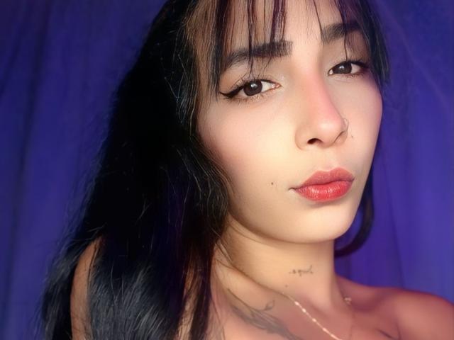 BrittanMelly with dark hair, bangs, and red lipstick, showing a nose ring and tattoo on her neck, with a subtle smile and makeup highlighting her eyes.