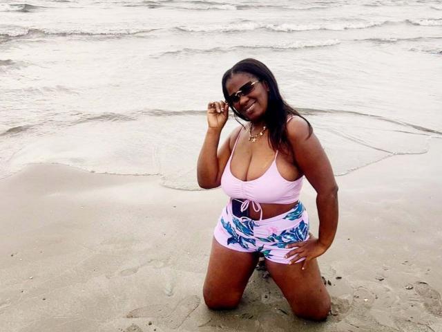 LinsayHott kneeling in sand, wearing pink top with big boobs, white shorts with blue floral, black sunglasses, smiling, right hand touching glasses, wet from small waves.