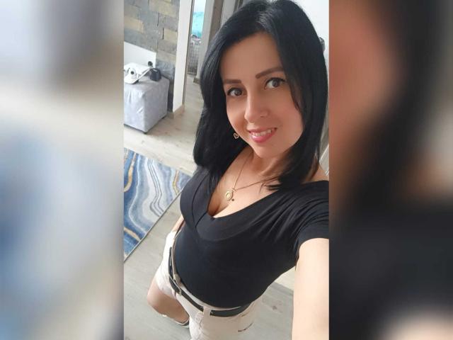 Briananice with black hair, smiling, wearing a black top, white pants, and a gold necklace, showing her small tits and a cute face. She's taking a selfie from above.