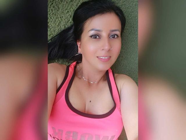 Briananice with medium tits, pink sports bra, black hair, nose ring, hoop earrings, subtle makeup, lying on green carpet, smiling, medium skin tone, small mole on chest.