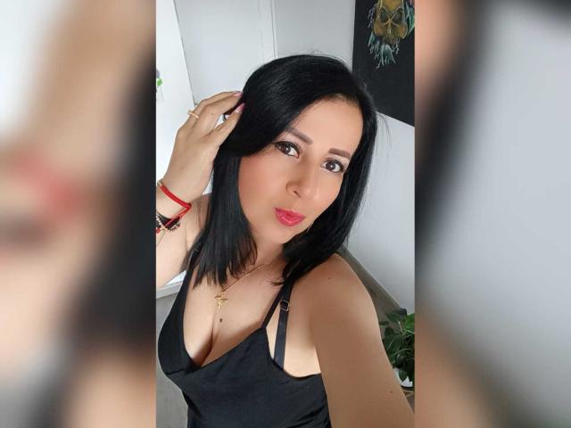 Briananice with black hair, red lipstick, and a black tank top, showing her tits, holding her hair, wearing a red bracelet, and a gold necklace.