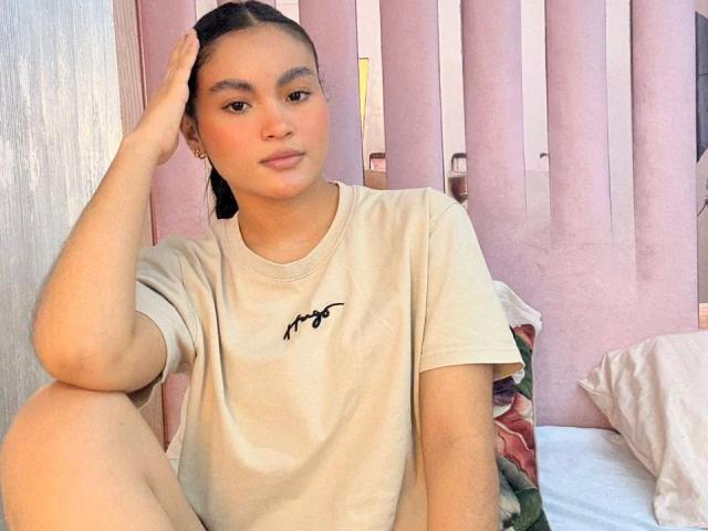 MiiaPalmer with medium brown skin, black hair in a ponytail, wearing a beige t-shirt, sitting with one knee up, hand on head, looking at camera, pink wall behind.