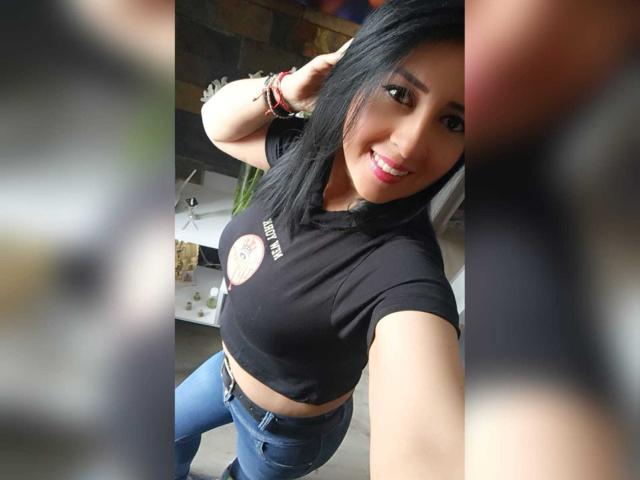 Briananice with black hair, smiling, wearing a black crop top showing her tits and blue jeans, taking a selfie, hand in hair, bracelets on wrist, pink lipstick.