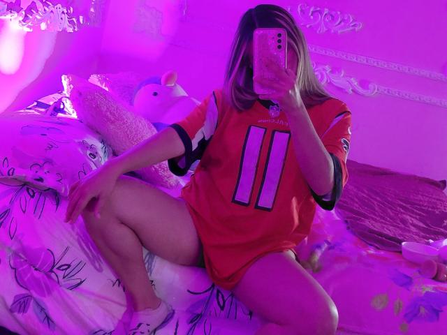 AngellVisen, sitting on a bed, wearing a red football jersey with number 11, taking a mirror selfie, showing her legs, with purple lighting, and stuffed toys around.