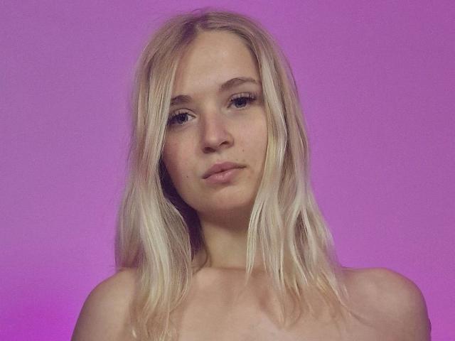 LillyOcean-hot has blonde hair, light skin, and a neutral expression with subtle makeup. She's topless, showing small tits and a small mole on her upper lip.