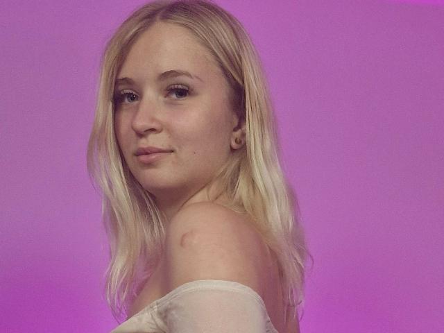 LillyOcean-hot with blonde hair, off-shoulder white top, light skin, small nose, subtle smile, and small ear piercings, set against a pink background.