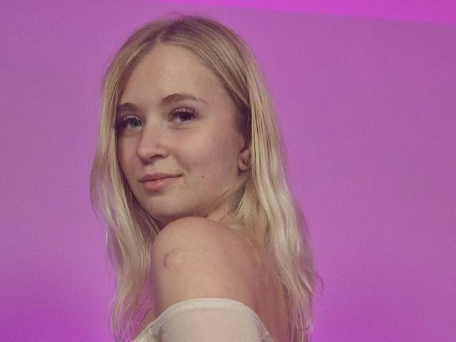 LillyOcean-hot with blonde hair, light skin, small tits, and a cute smile, looking over her shoulder, wearing an off-shoulder white top, against a pink background.