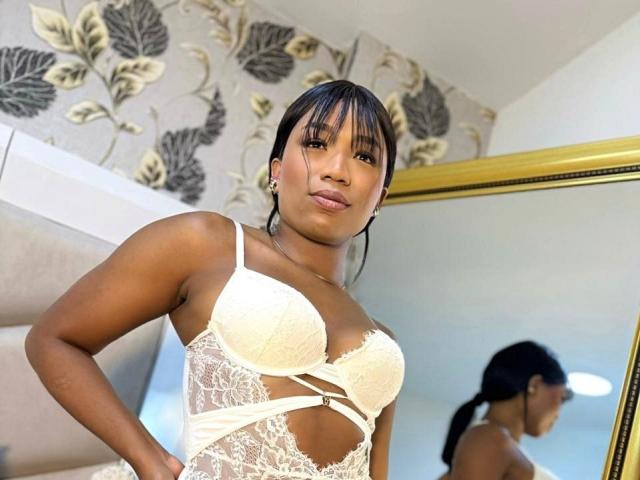 FarinaLyte, a dark-skinned woman with straight black hair and bangs, stands in a room. She wears a white lace bra and matching lingerie with cut-out sides. Her reflection is visible in a gold-framed mirror to the right. She has a confident expression and wears small earrings. The background features floral wallpaper. The lighting is bright, highlighting her smooth skin and the lace texture of her lingerie.