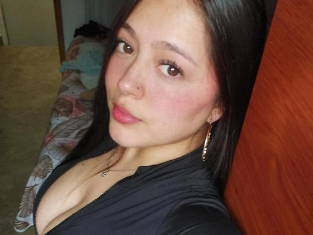 LiaJohnes has medium brown skin, long black hair, and small tits. She's wearing a black top with a deep cleavage, gold earrings, and a nose ring, looking at the camera.