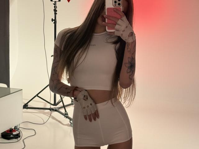 SensualMila with long brown hair, wearing a white sports bra and high-waisted shorts, fingerless gloves, tattoos on arms, taking a mirror selfie, red light in background.
