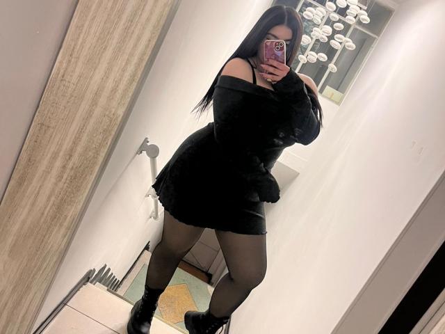 VenusxMusa with long black hair, wearing an off-shoulder black dress, black stockings, and black ankle boots, taking a mirror selfie, showing her curvy, voluptuous ass.