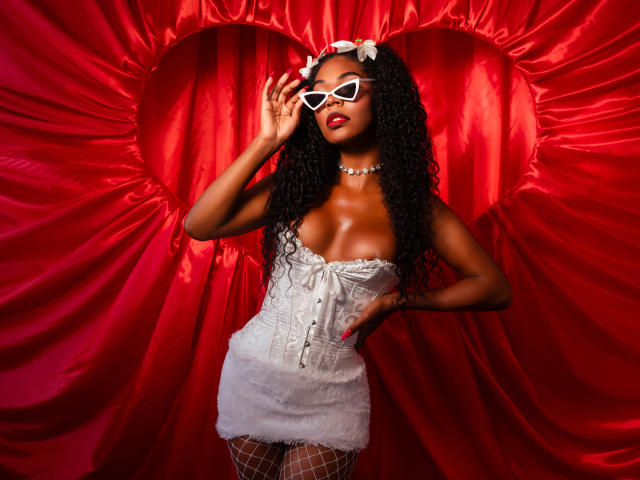 TianaParkerr with dark skin, curly black hair, white heart-shaped sunglasses, white corset, fluffy white mini-dress, fishnet stockings, white flower headpiece, pearl necklace, red background.