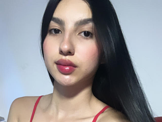 MilanMoon has long black hair, pink glossy lips, and wears a red spaghetti-strap top. She has a nose piercing and light skin with subtle pink blush on her cheeks.