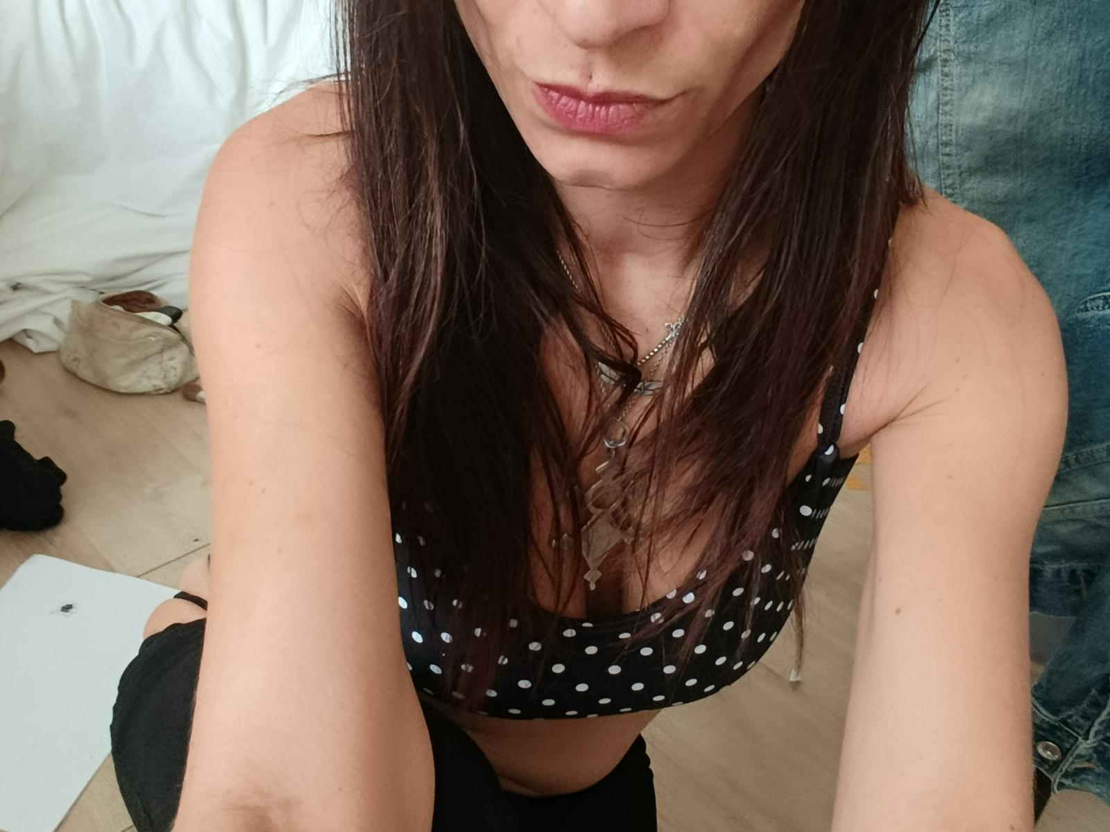 Lizoa with brown hair, pouty lips, wearing a black polka dot top, showing her small tits and cleavage, a necklace, and black pants, in a selfie.