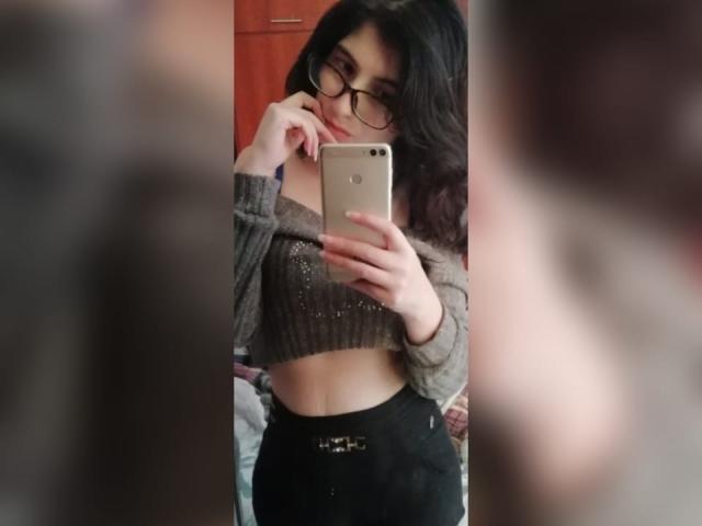 RubiDivine with dark wavy hair, glasses, grey crop top, black high-waisted pants, holding phone, taking mirror selfie, showing slim midriff, subtle makeup, soft light.