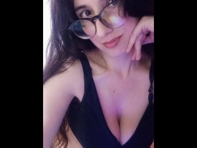 RubiDivine with glasses, black bra, showing her tits, hand resting on her cheek, long brown hair, pale skin, slight smile, pink lips, selfie, soft lighting.