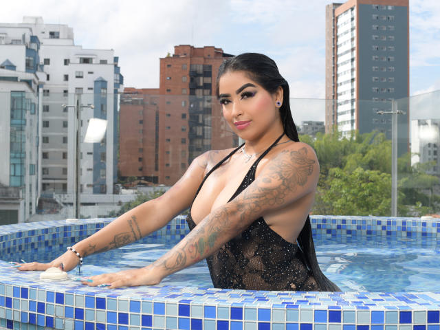 EmiilyRouss with tattoos, large boobs, in a blue-tiled hot tub, wearing a black, see-through, lace-up swimsuit, arms resting on the tub, black hair, pink lipstick, city background.
