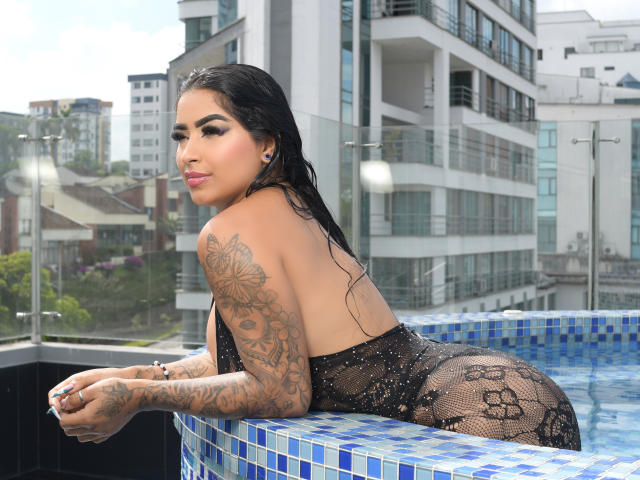 EmiilyRouss in a black, see-through lace bodysuit, wet black hair, tattooed arms, leaning over a blue-tiled pool, looking back, city buildings in the background.