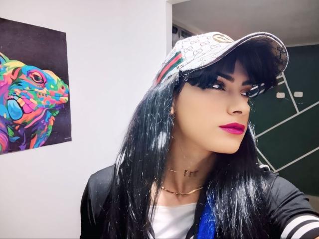 Sofia69xx with long black hair, wearing a white cap, black shirt, and pink lipstick, has a gold necklace and blue ribbon in her hair. Vibrant colorful art behind.