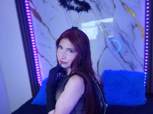 ScarletAss with red hair, black sheer top, black feather headpiece, looking over shoulder, blue pillow behind, purple LED strip lights, white and gray marble wall.