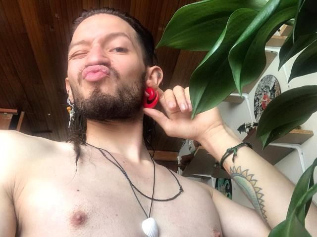 AngelDamianArt with a beard, puffy lips, and a red rose earring, shirtless, showing a tattoo on his right arm, and a white dog tag necklace.