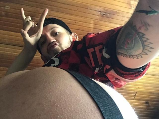 AngelDamianArt, shirtless, with tattooed arm, making peace sign, looking down at his bare belly and black underwear. Wood ceiling above. AngelDamianArt has short hair, beard, and a ring.