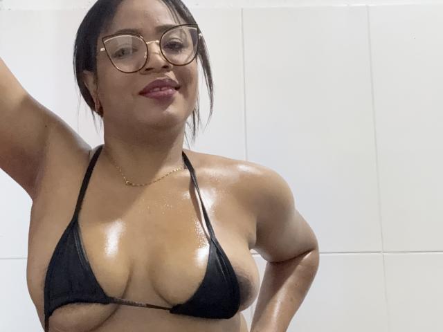 AliceBooty with wet skin, wearing a black bikini top, showcasing her large, shiny tits, glasses, and a gold necklace, smiling slightly.